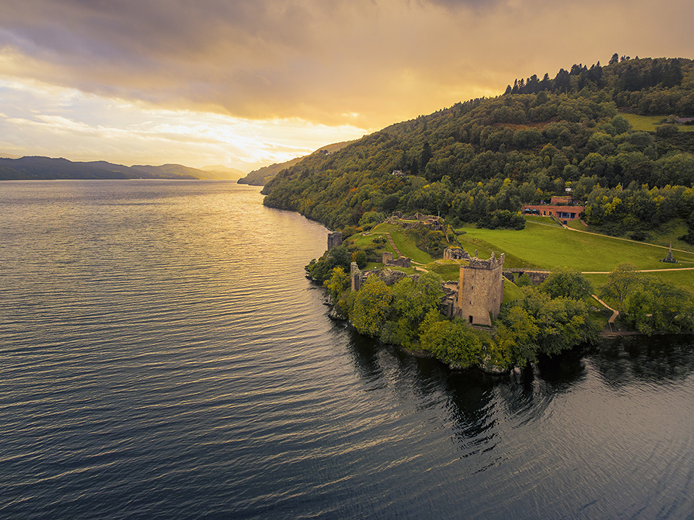 Castle in ruin on the shores of a loch, with the sun set in the background.